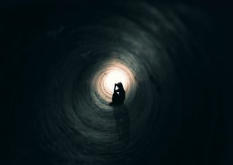 The silhouette of a praying woman sitting in a dark tunnel with a light at the end.